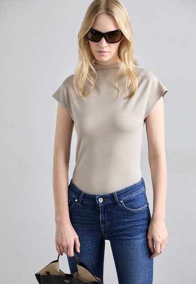 Top beige aderente con maniche corte arrotolate, abbinato a jeans blu scuro. La modella tiene una borsa nera. Sfondo neutro.