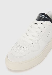 Hvit lær sneaker med perforert design, teksturerte semskede seksjoner, flate snøringer, og en svart lær hælflik med "Copenhagen Studios" merking.