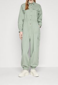 Mono verde claro de manga larga con cuello tipo solapa, cierre de botones en la parte delantera, cinturilla elástica y bolsillos laterales, combinado con zapatillas blancas.