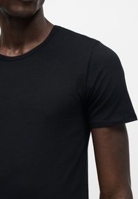 Mann trägt ein schlichtes schwarzes T-Shirt mit kurzen Ärmeln, dargestellt von den Schultern bis zur Brustmitte vor hellem Hintergrund.