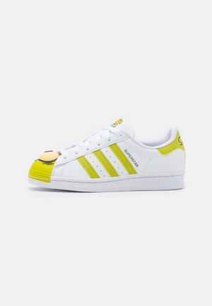 Biały sneaker Adidas Superstar z żółto-zielonymi paskami i językiem na pięcie, z postacią z kreskówki na nosku i języku.