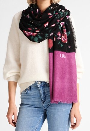 Femme portant un pull crème et un jean bleu, avec une grande écharpe noire et violette à motifs floraux drapée autour de son cou et son épaule.