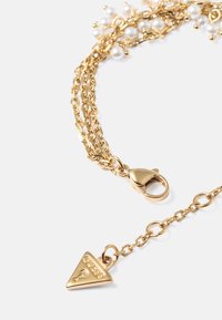Collana multi-strato in tono oro con perle sferiche, dotata di chiusura a granchio e un ciondolo triangolare contrassegnato "Guess".