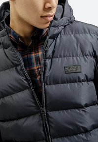 Granatowa puchowa kurtka z zamkiem z przodu i metką "CROSS JEANS"; pod nią znajduje się koszula w kratę w odcieniach czerwieni i niebieskiego.