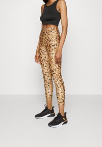 Donna che indossa un crop top senza maniche nero, leggings a vita alta marroni e neri con motivo, e sneakers Puma nere con accenti arancioni.