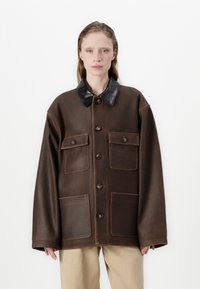 Brūna ādas jaka ar spīdīgu melnu apkakli, divām krūšu kabatām un divām apakšējām kabatām. Iespējas ietver lielas pogas un brīvu piegriezumu.