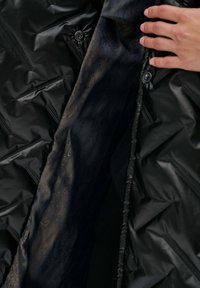 Zwarte puffy buitenstof met een gewatteerde textuur en zichtbare stiksels; interieur met donker marineblauwe stof met een gouden paisleypatroon en knoopdetails.