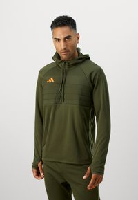 Felpa sportiva verde con un pannello frontale testurizzato, logo arancione e cappuccio regolabile. Il materiale sembra liscio con un design aderente e aperture per il pollice.