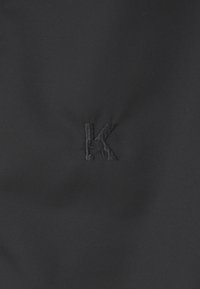 Schwarzer Stoff mit einer glatten Textur, der ein eingesticktes "K"-Logo in derselben Farbe zeigt, leicht versetzt.