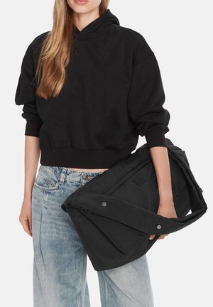 Žena v černé krátké mikině s kapucí a světle modrých džínách drží v náručí velkou černou prošívanou bundu.