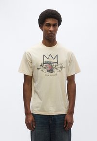 Εκρού βαμβακερό t-shirt με γραφικό σχέδιο κορώνας και μάσκας και κείμενο "KING ALPHONSO." Κοντά μανίκια, στρογγυλή λαιμόκοψη.