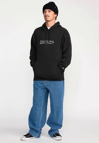 Zwarte hoodie met frontale afbeelding, kangoeroezak en capuchon met aantrekkoord; gecombineerd met lichtblauwe loszittende jeans en zwarte sneakers.