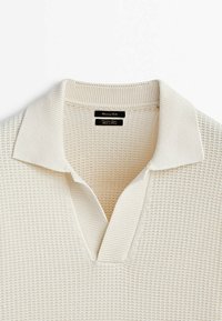 Vaalean beige lyhythihainen polo-paita, jossa on teksturoidun neuleen muotoilu, litteä kaulus ja v-aukkoinen pääntie. Brändilogo näkyy kauluksen sisäpuolella.