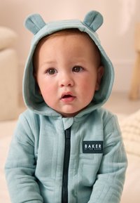 Lyseblå lynlåshættetrøje med bjørneører; quiltet tekstur; har et sort mærke med "BAKER by Ted Baker" branding på brystet.