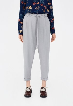 Mujer que lleva pantalones de cintura alta de color gris claro con cinturón de lazo, blusa floral de manga larga y zapatos marrón oscuro con cordones, de pie sobre un fondo blanco.