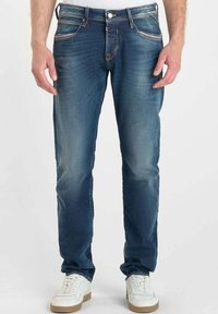 Jeans de mezclilla azul oscuro con un ajuste slim, con costuras en color naranja y un ligero desgarro. Combinados con zapatillas blancas para un contraste.