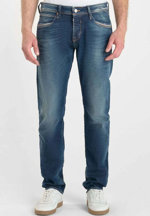 Jeans de mezclilla azul oscuro con un ajuste slim, con costuras en color naranja y un ligero desgarro. Combinados con zapatillas blancas para un contraste.