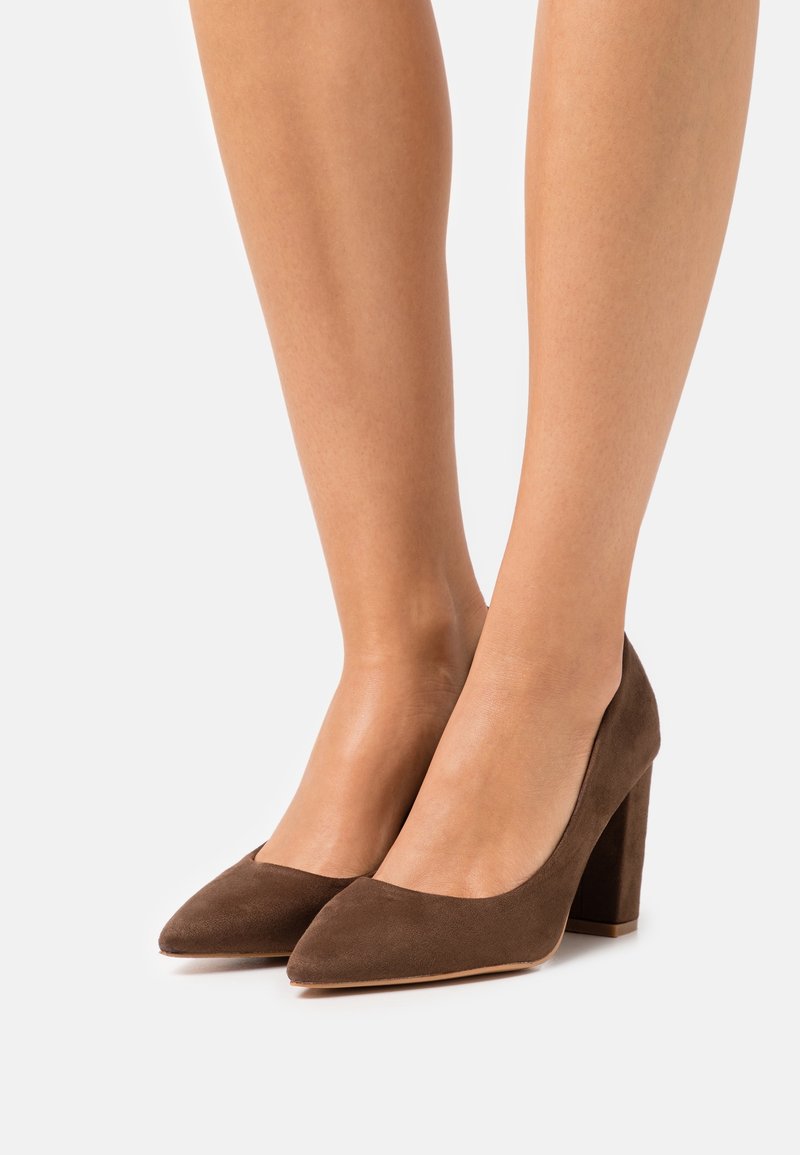 Escarpins en daim marron à talons hauts, bout pointu et talon épais. Texture lisse et design simple, sans motifs ni accents visibles.