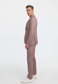 Világos rózsaszín öltöny, texturált anyagból, slim-fit szabással, sarkos hajtókákkal és két gombbal a bléné, fehér sneaker cipővel párosítva.