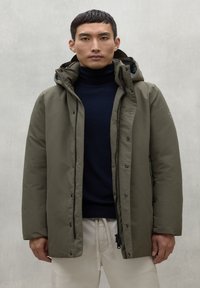 Parka acolchada verde olivo con capucha, cierre frontal de botones a presión y bolsillos laterales con cremallera, llevada sobre un cuello de tortuga azul marino y pantalones claros.