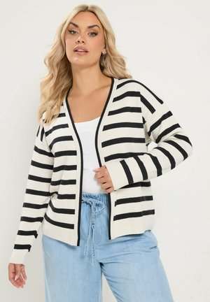 Frau mit langen blonden Haaren, die eine weiße und schwarz gestreifte Strickjacke, ein weißes Oberteil und hellblaue Hosen mit Kordelzug trägt.