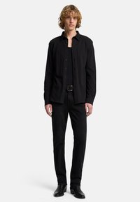 Schwarzes Hemd und schwarzes Tanktop übereinander getragen über schwarzen Jeans. Das Modell trägt einen schwarzen Gürtel und schwarze Stiefel. Einfaches Design ohne sichtbare Muster oder Akzente.