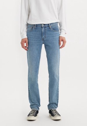 Homme portant un jean droit bleu clair, une chemise blanche à manches longues et des baskets noires avec des lacets blancs, debout sur un fond blanc.
