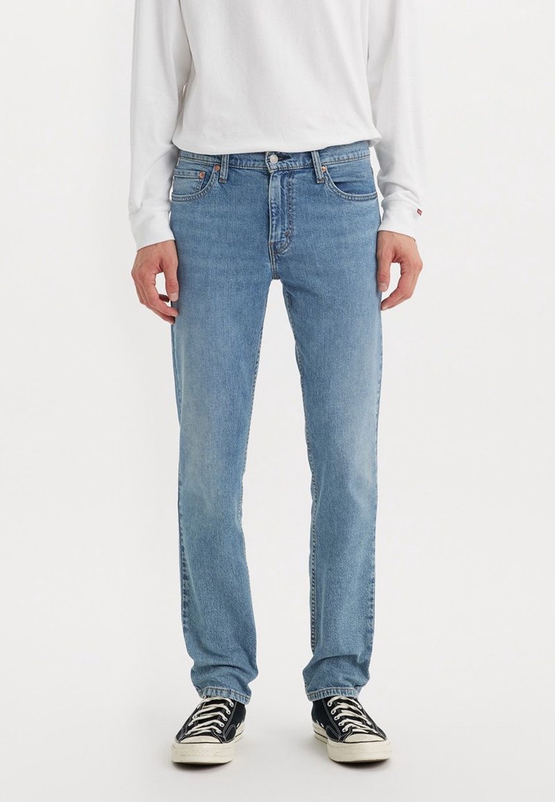 Homme portant un jean droit bleu clair, une chemise blanche à manches longues et des baskets noires avec des lacets blancs, debout sur un fond blanc.