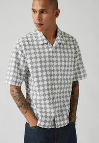 Camisa de manga corta en blanco con patrón geométrico negro. Cuenta con un cuello y cierre de botón, hecha de una tela ligera.