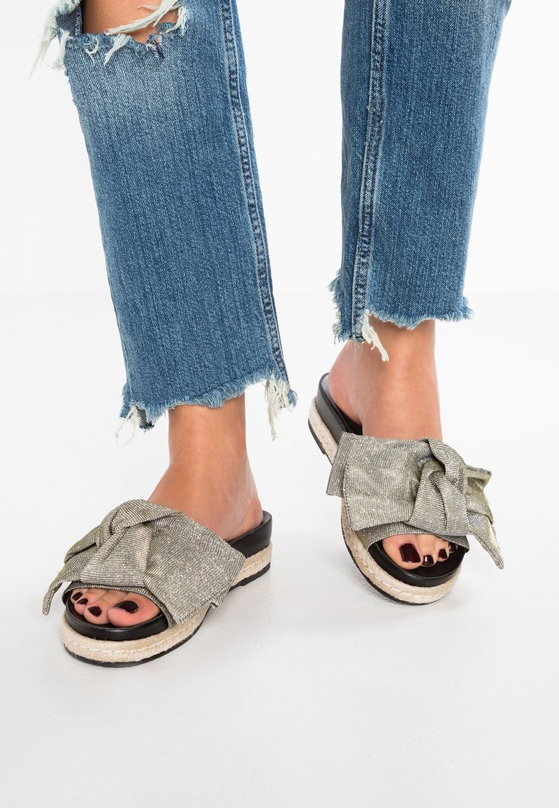 Pieds portant des sandales à bout ouvert avec de grands nœuds en tissu, associés à un jean bleu à ourlet effiloché, sur fond blanc.