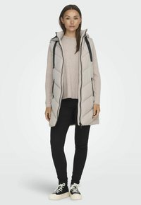 Gilet long matelassé gris clair avec fermeture éclair à l'avant, bretelles noires et motif en chevrons, porté sur un pull pâle et un pantalon noir.