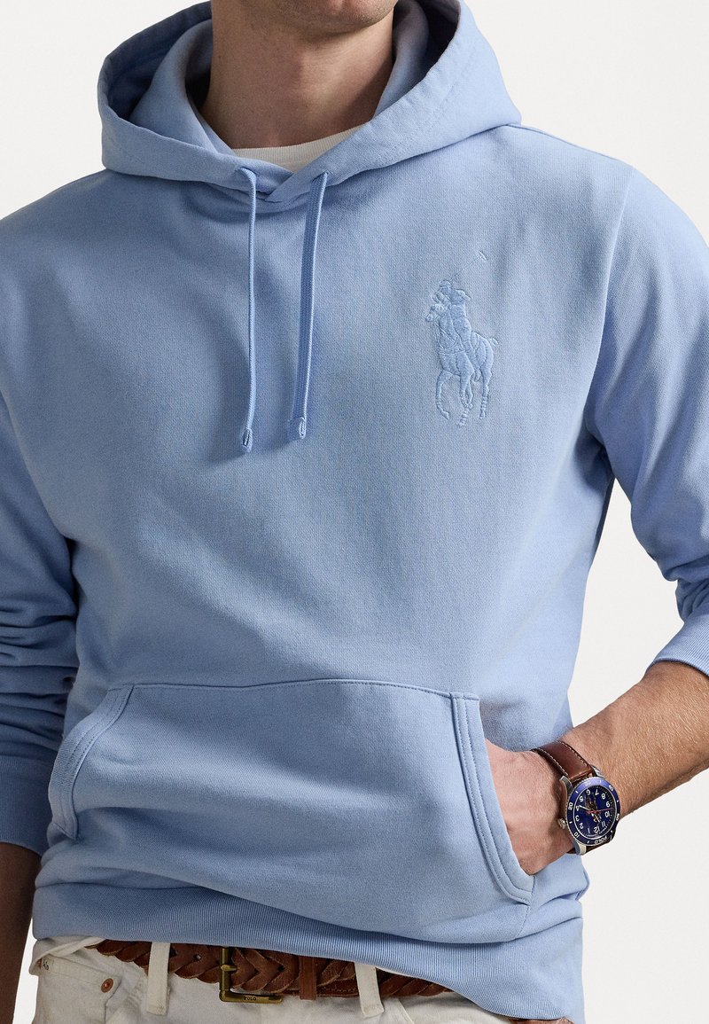 Sudadera azul clara hecha de un tejido suave, con un bolsillo canguro y un logo bordado en el pecho. Incluye cordones para ajuste.
