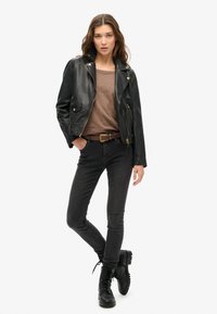 Vrouw in zwarte leren jas, bruine top, donkere skinny jeans, zwarte veterschoenen, staand met één hand in de zak op een witte achtergrond.