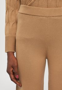 Strikket beige genser med kabelmønster og ribbestrikkede mansjetter, sammen med matchende ribbestrikkede bukser. Nærbilde som viser tekstur og passform.