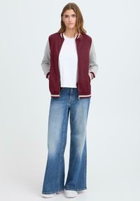 Femme debout avec les mains dans les poches, portant une veste de varsity bordeaux et grise, un t-shirt blanc, un jean large bleu et des baskets blanches.