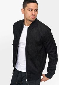 Schwarze Bomberjacke mit einer weichen, samtartigen Textur, ausgestattet mit einem Reißverschluss, gerippten Bündchen und Saum, getragen über einem weißen Hemd.