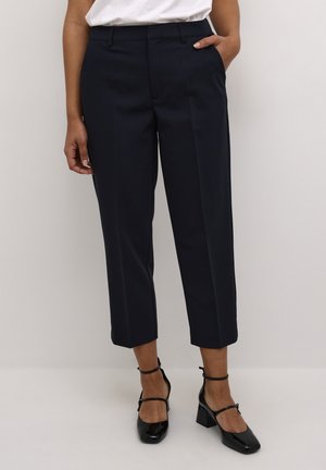 Navy tailored trousers with a straight fit, cropped length, and side pockets; paired with black patent heels and a white top.