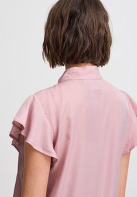 Blusa rosa chiaro a maniche corte con un leggero lucido, maniche arricciate e un colletto semplice, che mette in mostra una texture del tessuto liscia.