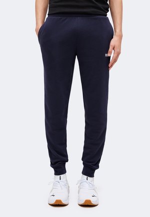 Persoană purtând pantaloni sport Puma bleumarin cu buzunare laterale și adidași albi Puma, stând în fața unui fundal simplu și deschis.