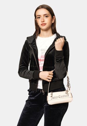 Jonge vrouw draagt een zwarte fluwelen hoodie met rits en broek, wit grafisch T-shirt, houdt een beige Juicy Couture handtas om haar pols.