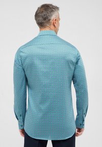 Een langemouwen shirt van turquoise stof met een geometrisch patroon van kleine vierkanten in paars en groen. Voorzien van knoopmanchetten en een spread collar.