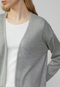 Hellgrauer Cardigan aus weichem Strickmaterial, mit V-Ausschnitt und offenem Vorderteil. Tragen über einem weißen, gerippten Shirt mit lockeren Ärmeln.