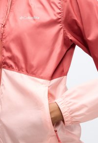 Ροζ μπουφάν Columbia δίχρωμο με ελαστικό μανσέτα και χέρι τοποθετημένο στην μπροστινή τσέπη, που δείχνει την υφή του υφάσματος και τις λεπτομέρειες των ραφών.