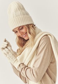 Smilende kvinde iført en cremefarvet strikket hat, matchende handsker og tørklæde over en beige sweater, der justerer en handske på sin hånd.
