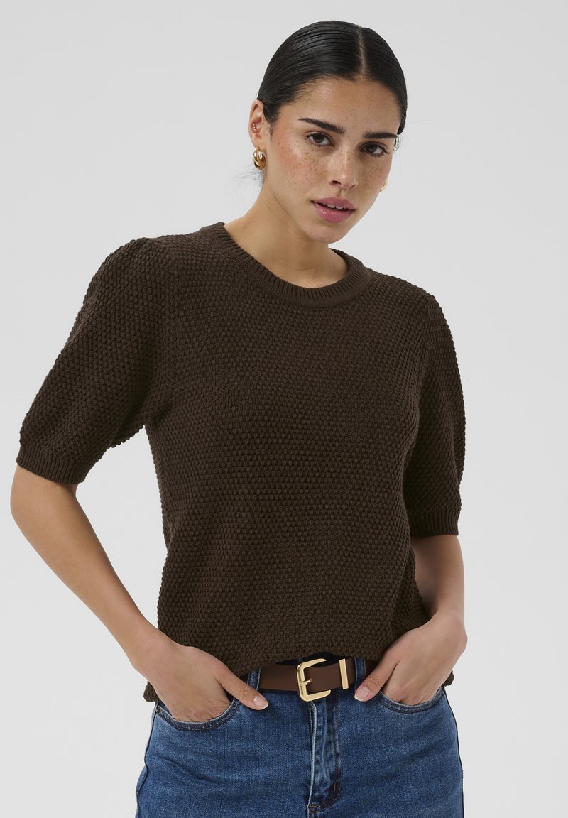 Femme aux cheveux foncés portant un pull marron texturé à manches courtes, un jean bleu et des boucles d'oreilles créoles dorées, debout les mains dans les poches.