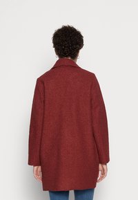 Röd texturerad kappa med urklippt krage, avslappnad passform och långa ärmar. Har en enfärgad design. Bärs över blå jeans.