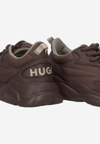 Brown athletic shoes with a textured fabric upper, featuring a contrasting beige collar and laces. The back has a prominent "HUGO" logo.