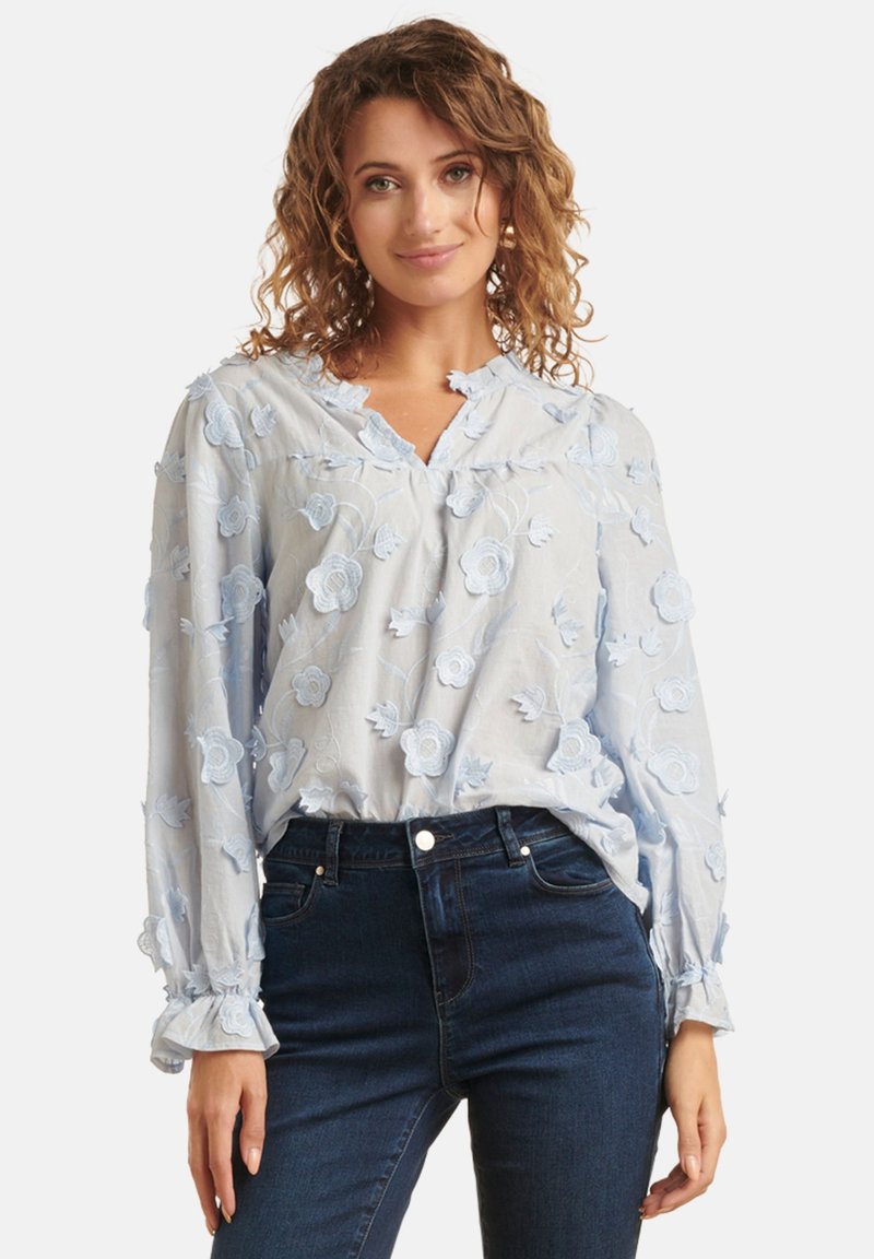Light blue blouse with floral embroidery, v-neckline, long sleeves with ruffled cuffs, paired with dark blue jeans. Soft texture.