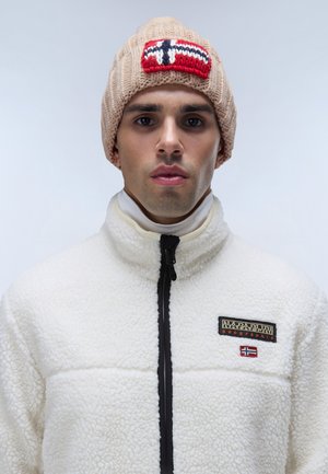 Beige gebreide beanie met een rood, wit en blauw geborduurd label; witte fleece jas met een zwarte rits en een contrasterend logo label.