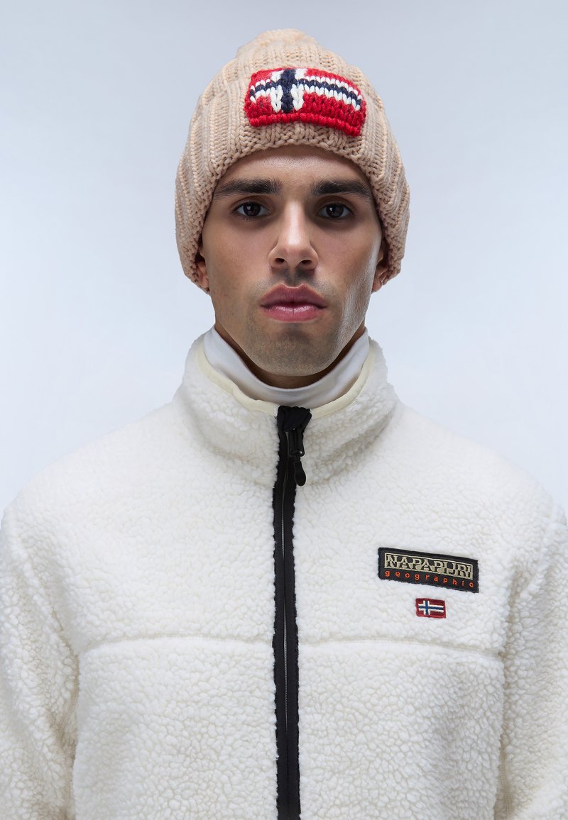 Beige gebreide beanie met een rood, wit en blauw geborduurd label; witte fleece jas met een zwarte rits en een contrasterend logo label.